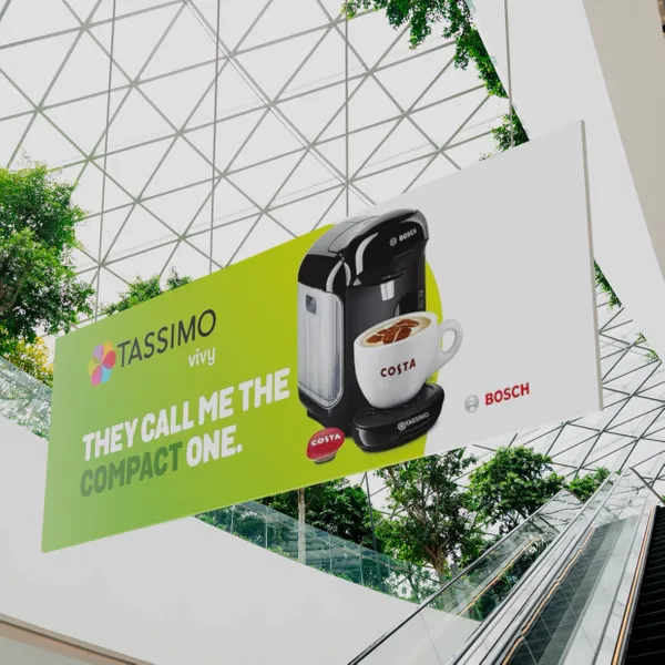 A coffee machine ad reading 'TASSIMO vivy – THEY CALL ME THE COMPACT ONE' displayed above an escalator in a contemporary setting with a geometric glass ceiling.